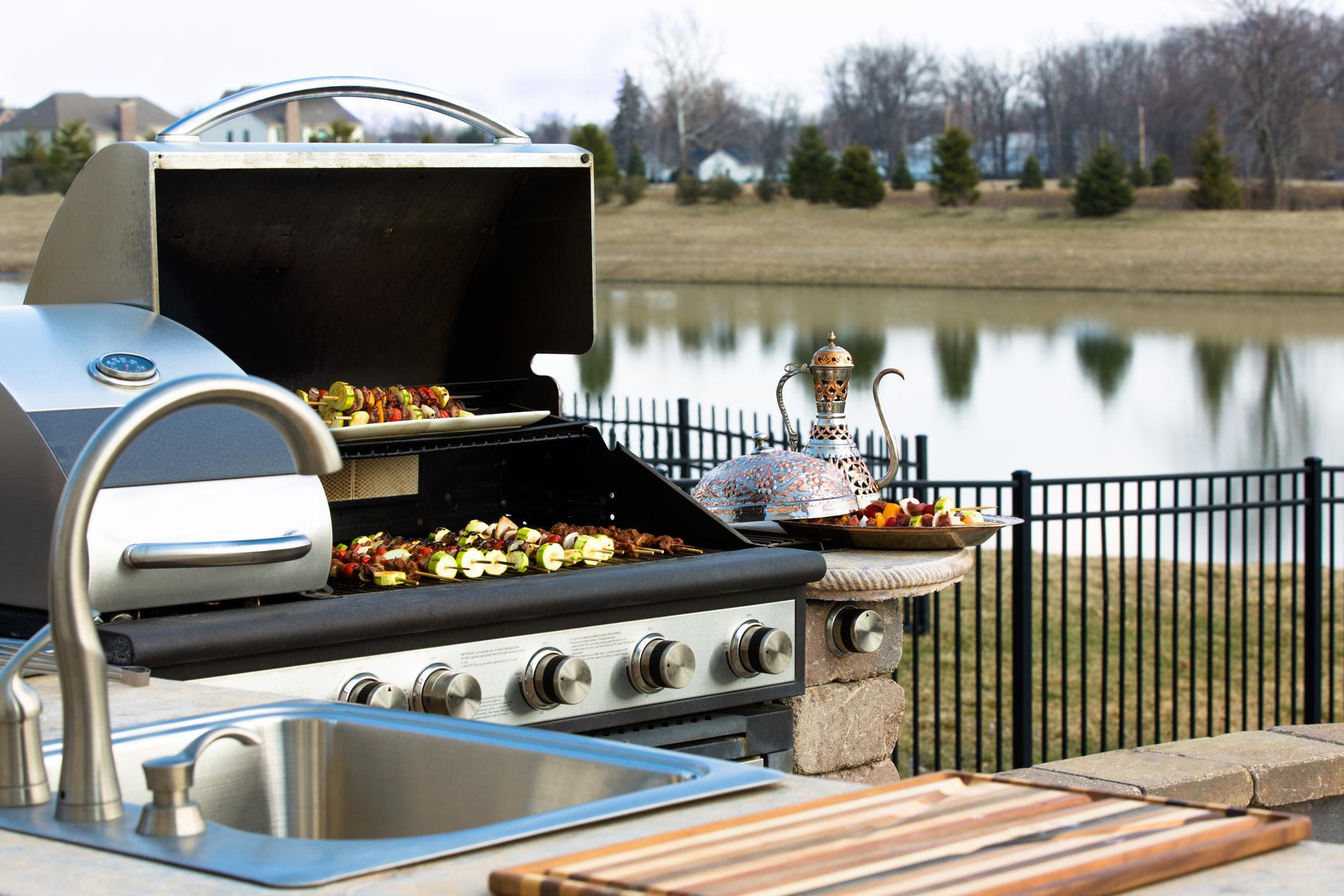 Built-in stainless gas braai with its hood open and kebabs on the grill, beside an outdoor sink overlooking a dam