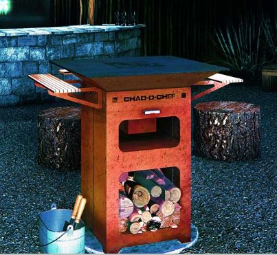 Chad-O-Chef Boma Braai in corten steel at night with logs stacked in the base and an ice bucket beside it