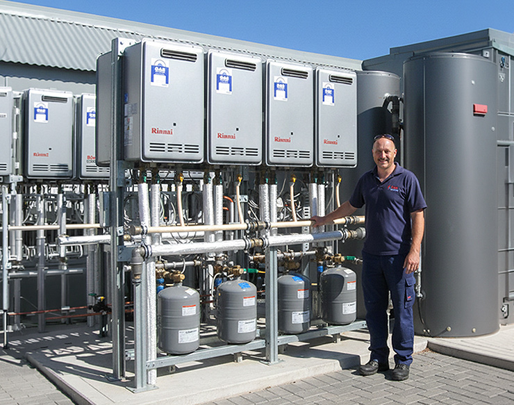 Bank of Rinnai gas water heaters mounted outdoors on a steel frame, with insulated pipework and expansion vessels below and a technician standing alongside